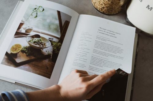 From above of crop faceless woman reviewing article in recipe book in kitchen